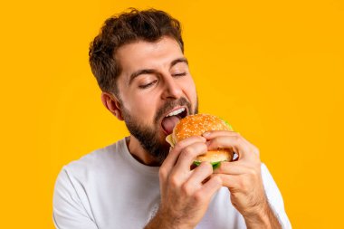 Abur cubur yiyen. Aç bir adamın lezzetli bir hamburgerin tadını çıkarması, sarı stüdyo arka planında paket burgerden bir ısırık alması. Diyet ve aşerme arasındaki beslenme dengesi