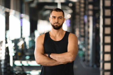 Spor salonunda kolları çapraz duran, kendine güvenen kaslı bir adam. Arka planda spor kıyafetleri olan yakışıklı erkek sporcu eğitime hazır, kameraya bakıyor, serbest alan.