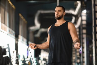 Ellerinde ip atlayan, spor salonunda duran, kardiyo antrenmanına hazırlanan yakışıklı kaslı, motive olmuş, fotokopi alanından uzağa bakan erkek sporcunun portresi.