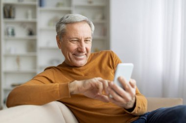 Evdeki kanepede dinlenen mutlu, gri saçlı, yaşlı adam akıllı telefon, web sörfü, kaydırma, güzel mobil uygulama, kopyalama alanı kullanıyor. Yaşlılar ve teknoloji