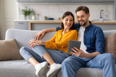 Neşeli genç çift koltukta rahatça oturuyor, dijital tableti paylaşıyor, kadın öne eğiliyor ve adam aleti tutuyor, ikisi de mutlu görünüyor, internette sörf yapıyor.