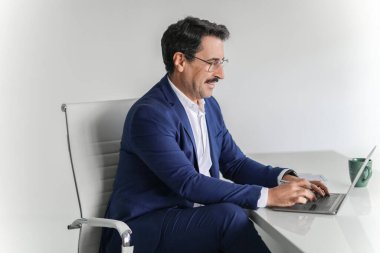 Odaklanmış, mavi takım elbiseli, olgun, beyaz bir işadamı modern çalışma alanındaki dizüstü bilgisayarında işine dalmış bir fincan kahveyle yazı yazıyor. İş, aletle iş.
