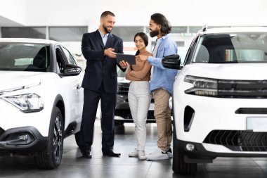 Dijital tabletli çekici araba satıcısı, bir bayilik salonunun cilalı arabaları arasında, özenli bir genç doğu çiftiyle yeni bir arabanın ayrıntılarını paylaşıyor.