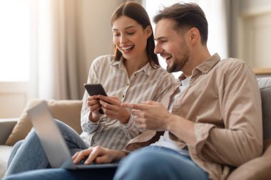 Neşeli genç bir kadın dizüstü bilgisayarla birden fazla iş yapan gülen bir adama akıllı telefon gösteriyor. İkisi de evdeki kanepede keyifli anlar yaşıyor.