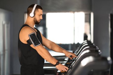Kulaklıklı ve kol bandı takan genç bir sporcu koşu bandındaki ayarları ayarlıyor, kaslı adam iyi donanımlı bir spor salonunda kardiyo seansına hazırlanıyor.
