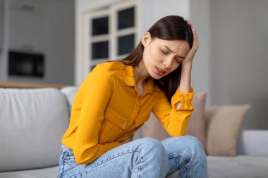 Sıkıntılı genç bir kadın kanepeye oturur, acı içinde başını tutar, şiddetli baş ağrısı ve mide ağrısını gösteren acı dolu bir ifadeyle, kendini kötü hisseder.