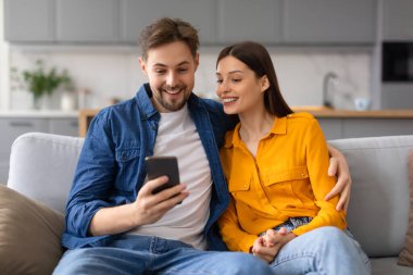 Neşeli eşler kanepeye yakın otururlar, akıllı telefona bakarken neşeli anları paylaşırlar, mutluluk ve beraberlik ifadeleriyle.