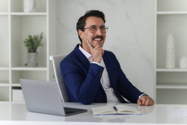 Mavi takım elbiseli düşünceli beyaz işadamı elinde dizüstü bilgisayar ve defterle masasında oturuyor, elleri çenesinde, şık bir ofiste dalgın dalgın bakıyor, düşün. İş, iş planlaması