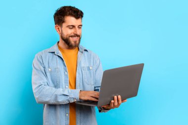 Mutlu sakallı genç adam dizüstü bilgisayarında yazı yazıyor. Stüdyoda mavi arka planda web sörfü yapıyor. Çevrimiçi işlere dalmış serbest çalışan adamın portresi. E-öğrenme ve uzaklık işi