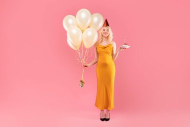 Uzun sarı cüppeli ve parti şapkalı zarif sarışın, bir elinde doğum günü pastası, diğer elinde krema balonları, pembe arka planda poz veriyor.