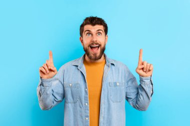 Pozitif genç beyaz adam yukarı doğru işaret ediyor. Stüdyo ortamında mavi arka planlı bir satış önerisi. Heyecanlı adamın portresi kafasının üstünde bir şey gösteriyor.