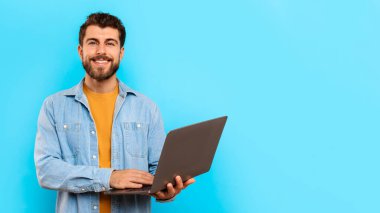 Serbest, online iş. Dizüstü bilgisayarda çalışan mutlu sakallı adam, mavi stüdyo geçmişinde cihazla poz veriyor, internet reklamları yapıyor, dijital ürünleri tanıtıyor. Panorama, boşluğu kopyala