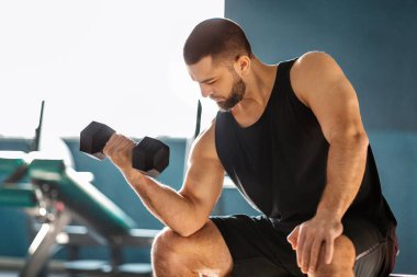 Genç erkek sporcu, Dumbbell ile antrenman yapıyor, Modern Spor Salonu 'nda egzersiz yaparken bikini kıvırıyor, Kol kaslarını çalıştırıyor, vücut geliştirmenin keyfini çıkarıyor, bedava alan yaratıyor.