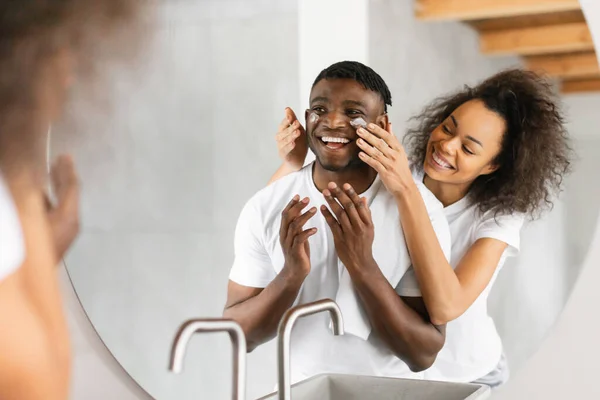 Black young spouses sharing playful skincare routine at home bathroom, wife applying moisturizer on husbands cheeks standing behind him near mirror. Family Skin Care Concept