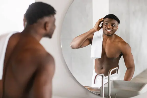 Handsome Young African American Man Engages In Morning Selfcare, Standing Shirtless In Modern Bathroom, Smiles At His Reflection In Mirror Touching Hair, Enjoying Daily Routine Of Grooming