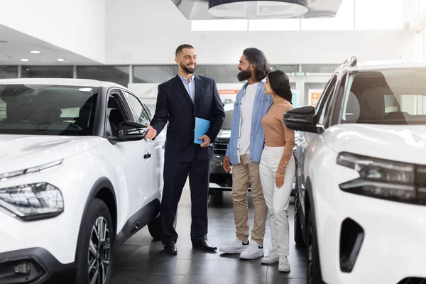 Car purchase or rental. Positive young indian spouses speaking to salesman about buying new auto at dealership. Successful millennial manager showing clients choice of vehicles at salon