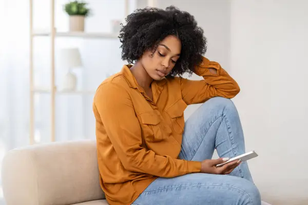 Pensive sad African American woman with curly hair, wearing a mustard blouse, sitting on a couch looking at her phone with a contemplative expression, chatting, read news