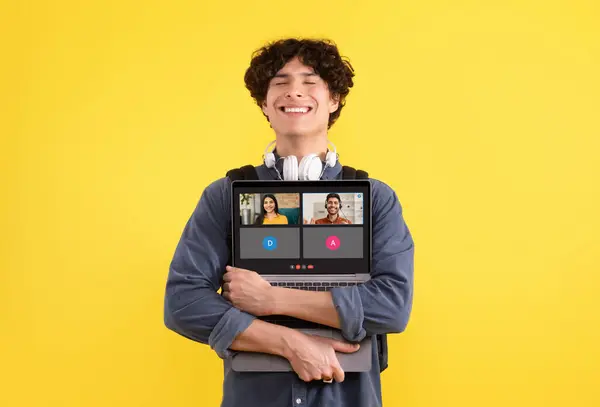 一位脖子上挂着卷发和耳机的年轻人 一边心满意足地微笑着 一边抱着笔记本电脑 在黄色的背景上播放着一个活跃的视频通话 社交网络 — 图库照片