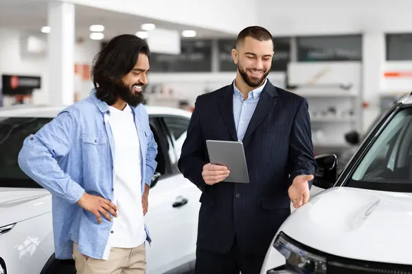 Cheerful indian guy standing by nice white car, talking to dealer holding digital tablet. Wealthy eastern young man having conversation with handsome sales manager at luxury showroom