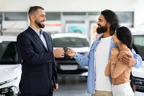 Car purchase or lease concept. Cheerful young indian couple taking car key from auto salesman at modern dealership. Millennial eastern spouses buying new automobile at showroom store