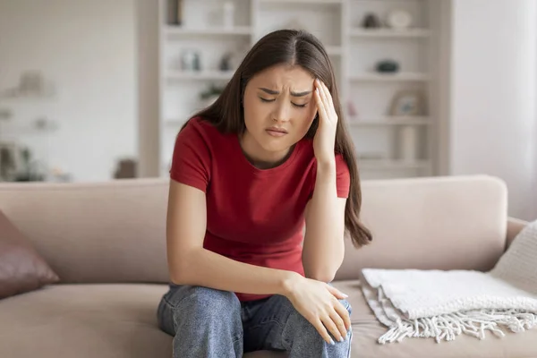 Anxious young asian woman with hand to her head suffering signs of headache or stress, depressed korean lady sitting on couch in living room interior, millennial female feeling unwell at home