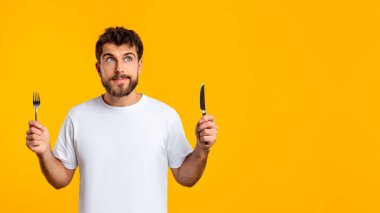 Aç sakallı Avrupalı adam elinde bıçak ve çatal ile restoran yemeğinin tadını çıkarmaya hazır bir şekilde stüdyoda sarı arka planda bekliyor. Açlık ve yemek aşermeleri. Boşluklu Panorama