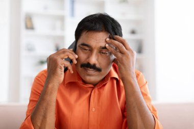 Turuncu tişörtlü, bıyıklı olgun Hintli adam oturma odasındaki koltukta oturuyor, evde telefon görüşmesi yapıyor, kötü haberler duyuyor, kafa kafaya dokunuyor, yakın plan yapıyor.