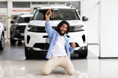 Duygusal yakışıklı doğu çocuğu dizlerinin üzerinde duruyor ve elini kaldırıyor yepyeni beyaz arabasının anahtarını gösteriyor, kopyalama alanı. Zengin Hintli adam lüks galeriden araba alıyor.