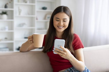 Güzel Asyalı genç bir kadın akıllı telefon ve kahveyle koltukta dinleniyor, cep telefonu kullanan Koreli bir kadın gülümsüyor ve evde dinlenirken sıcak içkinin tadını çıkarıyor.