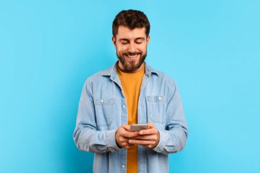 Mutlu genç adam akıllı telefonuyla bir uygulamayı tarıyor, stüdyoda mavi arka plana karşı duruyor, online iletişim ve mesajlaşma ile meşgul, milenyum adamı modern teknoloji sunuyor.