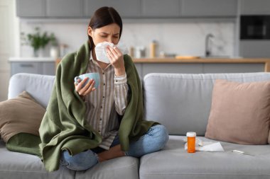 Hasta genç bir kadın battaniyeye sarılmış, burnunu siliyor ve elinde sıcak bir içecek, masada ilaç, soğuk algınlığı ya da evde grip olduğunu gösteren bir bardak tutuyor.