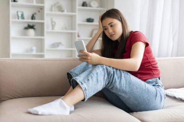 Akıllı telefonlu yalnız bir Asyalı kadın evdeki kanepede oturuyor, cep telefonu ekranına bakıp mesaj bekleyen, depresyon geçiren, oturma odasında dinlenen Koreli genç bir bayanı üzüyor.