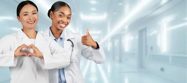 Two happy asian, african american millennial healthcare professionals in lab coats in a hospital, one making a heart gesture with her hands and the other giving a thumbs-up