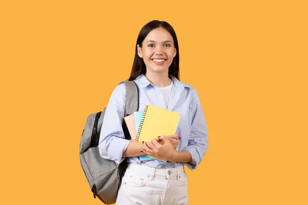 青いシャツのハッピーヨーロッパの女性学生はノートを保持し 灰色のバックパックを着て 明白な黄色の背景に対してカメラで立っています — ストック写真