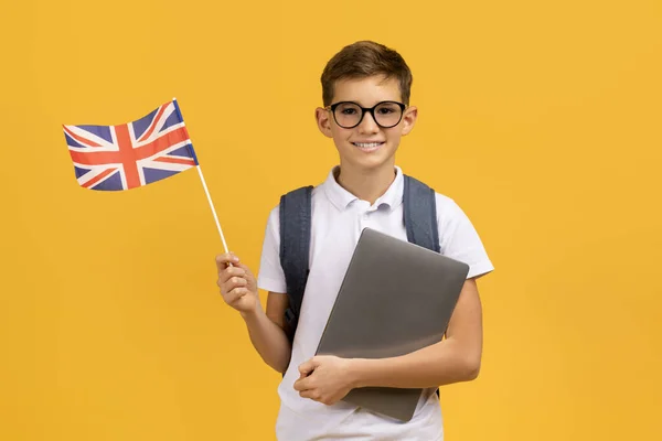 留学について 笑顔の学校のボーイ ラップトップとイギリスのフラグを保持し ハッピーティーン男性の子供は バックパックを身に着けています オンライン外国語コースをお勧め 黄色の背景に立っています — ストック写真