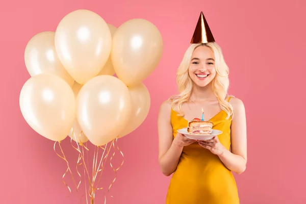 黄色いドレスとお祝いの帽子のラディアントブロンドの女性 黄金の風船の横のケーキのプレートで微笑んで ピンクのスタジオ背景でお祝い — ストック写真