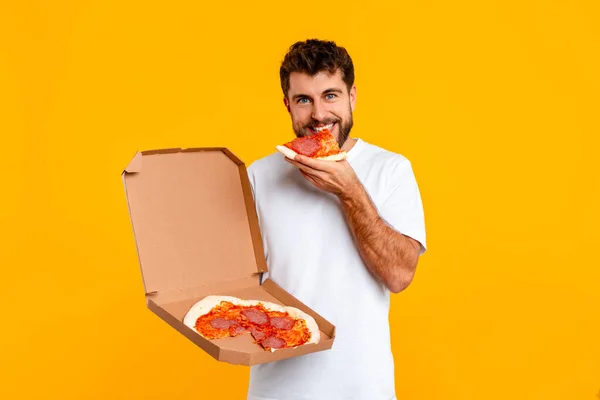 Hungry young European man bites into slice of pizza, emphasizing concept of comfort eating and junk food consumption, standing on yellow background, looking at camera. Nutrition habits