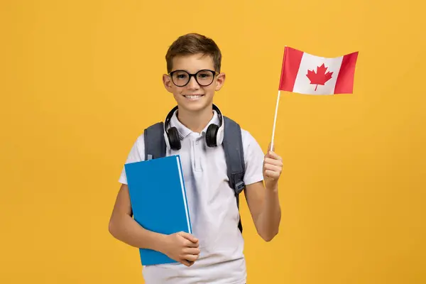 インターナショナル教育 カナダの旗とワークブックを保持しているハッピーティーンボーイ イエロースタジオの背景にポーズ笑顔の学校のボーイの肖像 海外留学を楽しむ コピースペース — ストック写真