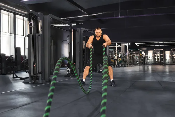 Crossfit training. Motivated muscular man exercising with battle ropes at gym, young caucasian male athlete engaging in high-intensity workout, challenging his strength and endurance in sport club