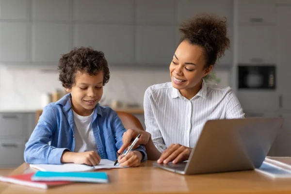 Smiling young black mother assists her curly-haired son with his schoolwork using laptop at wooden kitchen table, capturing moment of learning