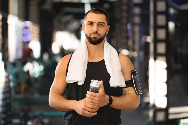 Muscular young man standing in gym with towel draped over his shoulders and water bottle in hands, handsome caucasian male athlete exuding confidence after his workout routine, copy space