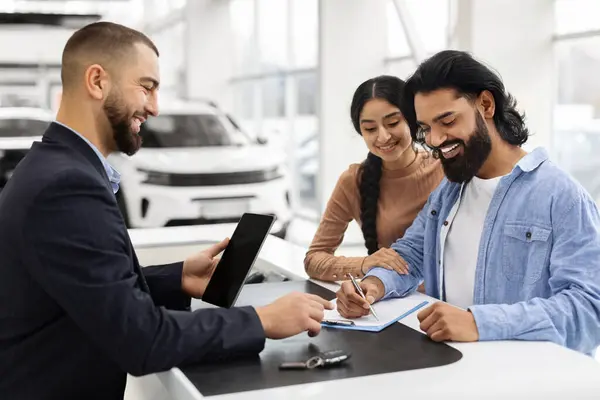 Wealthy millennial indian family buying new car in showroom, standing by counter with handsome man sales assistant in suit, filling papers and having conversation. Car retail, leasing, auto showroom
