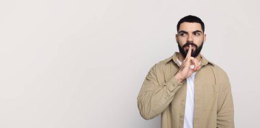 Ciddi, yakışıklı, sakallı, orta doğulu genç bir adam. Parmak dudaklı, sessiz bir jest yapıyor. Boş alana bakıyor. Haber ve dedikodu jesti