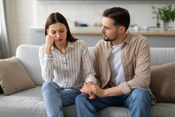 stock image Compassionate young man holding hand of worried woman, offering comfort while they sitting together on comfy gray sofa in well-lit living room