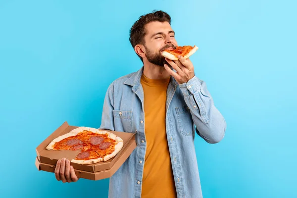 Hungry millennial man enjoys taste of pizza slice holding opened delivery box over blue studio backdrop, Concept of overeating and the pleasure from cheat meal during diet