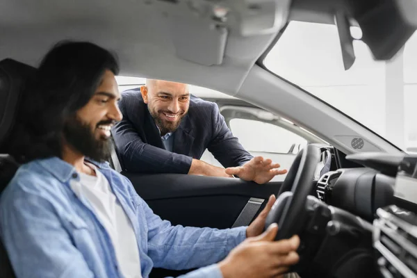 Happy eastern man sitting in white luxury car listening to sales manager and smiling. Closeup of bearded indian guy buying brand new comfortable auto in showroom, side view