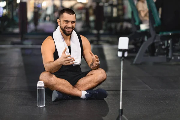 Blogging Concept. Smiling Muscular Man Recording Video Content At Gym, Handsome Sporty Bodybuilder Couch Talking At Smartphone Camera While Sitting On Floor In Modern Fitness Club, Copy Space