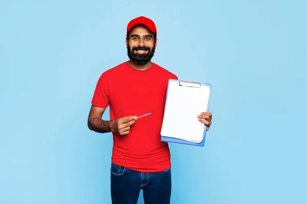 Cheerful indian delivery man in red uniform holding pen and clipboard with blank paper, ready for signature, standing against vivid blue background