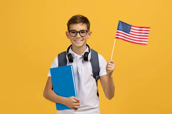 快乐的少男学生 拿着印有美国国旗的笔记本 快乐的少男学生在国外学习英语 看着镜头笑着 在黄色的工作室背景上摆姿势 复制空间 — 图库照片