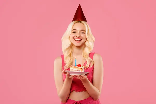 光栄な若いブロンドの女性はパーティー帽子をかぶり 単一のろうそくで誕生日ケーキを開催し 彼女の顔は活気に満ちたピンクの背景に対して喜びで点灯しました — ストック写真